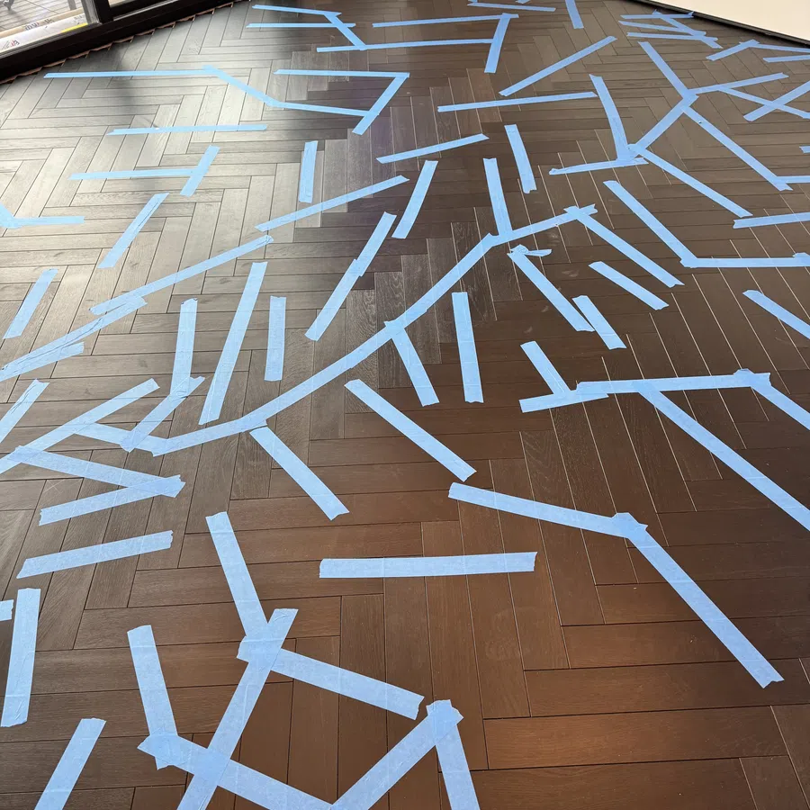 Dark hardwood floor with blue painter's tape arranged in random patterns during home renovation near sliding glass doors.