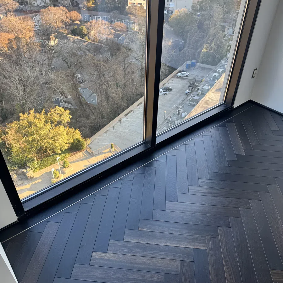 Dark herringbone wood floor by large window with cityscape and trees outside during sunset light.