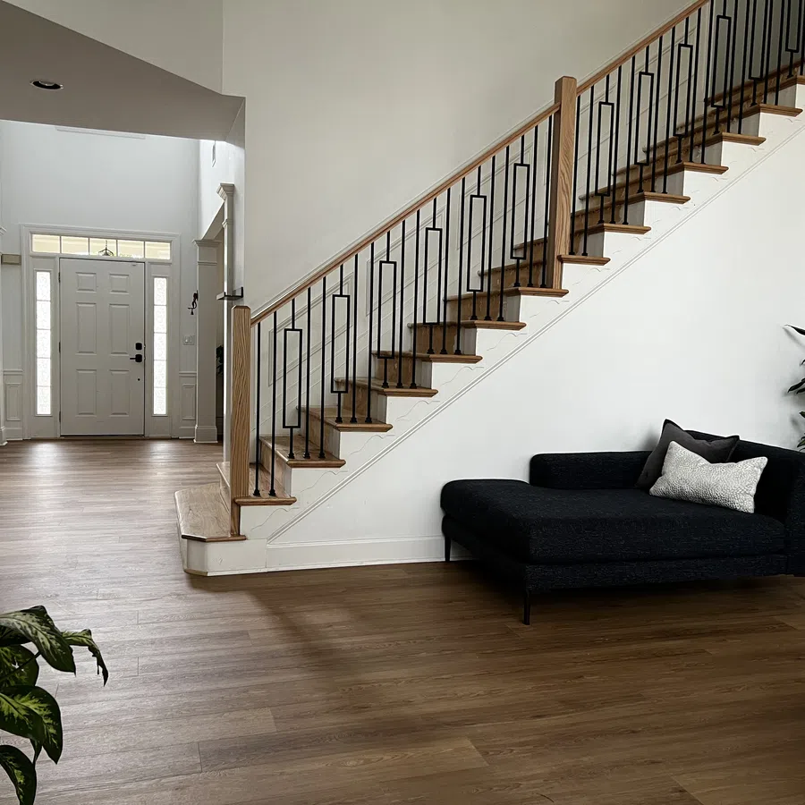 Modern entryway with wooden staircase, black chaise lounge, potted plants, and light hardwood floors in a bright home.