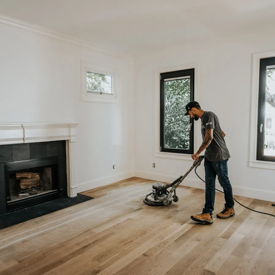 Man using a floor polishing machine to clean hardwood floors in a bright, empty living room with fireplace.