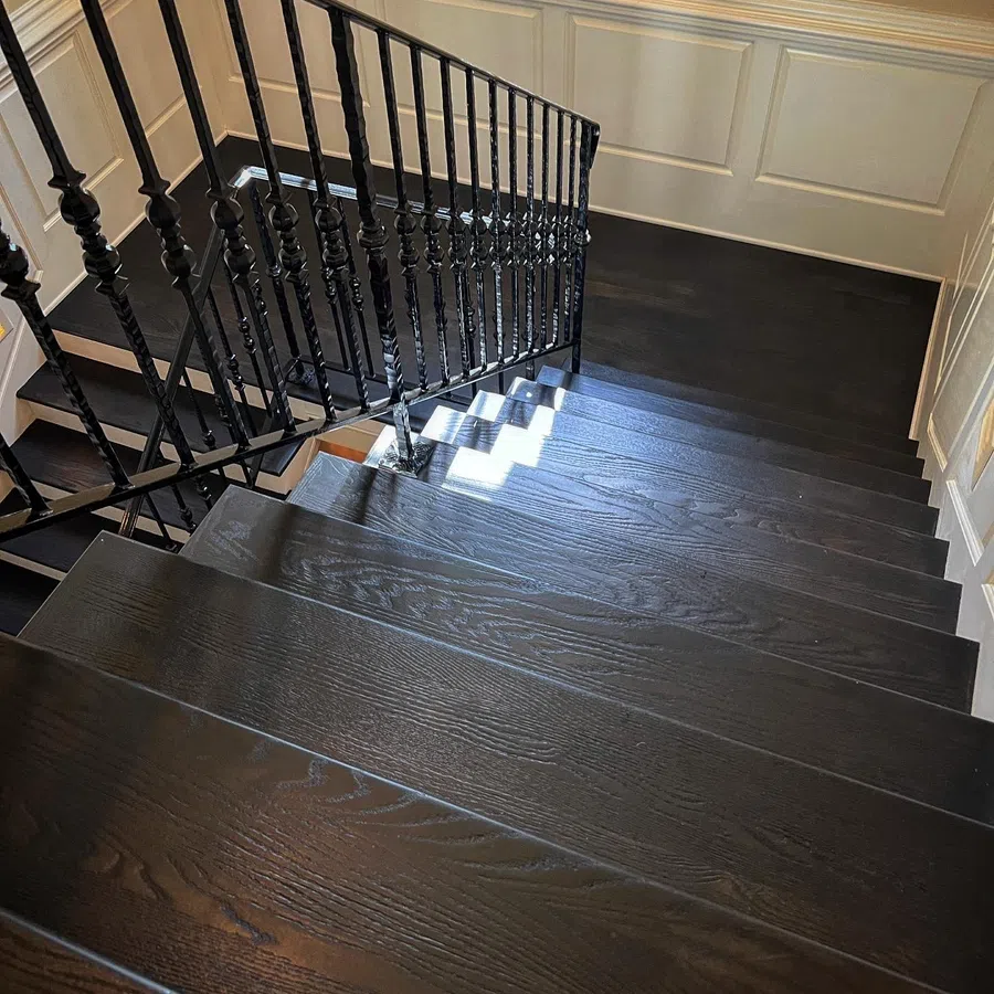 Dark wooden staircase with decorative black metal railing and white paneled wall in bright interior.