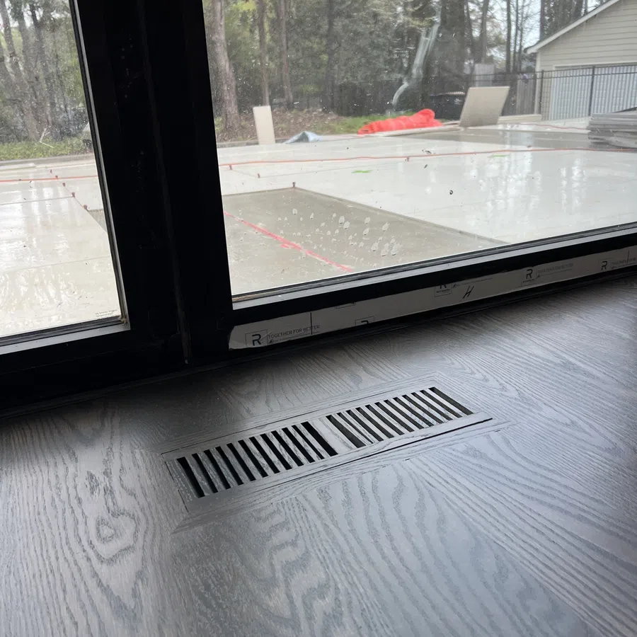 Wood floor vent near sliding glass door with outdoor patio and trees visible through the glass.
