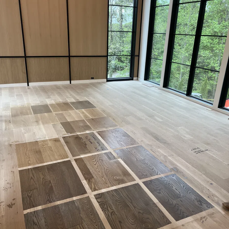 Sample wooden flooring swatches arranged on light wood floor in bright room with large black-framed windows