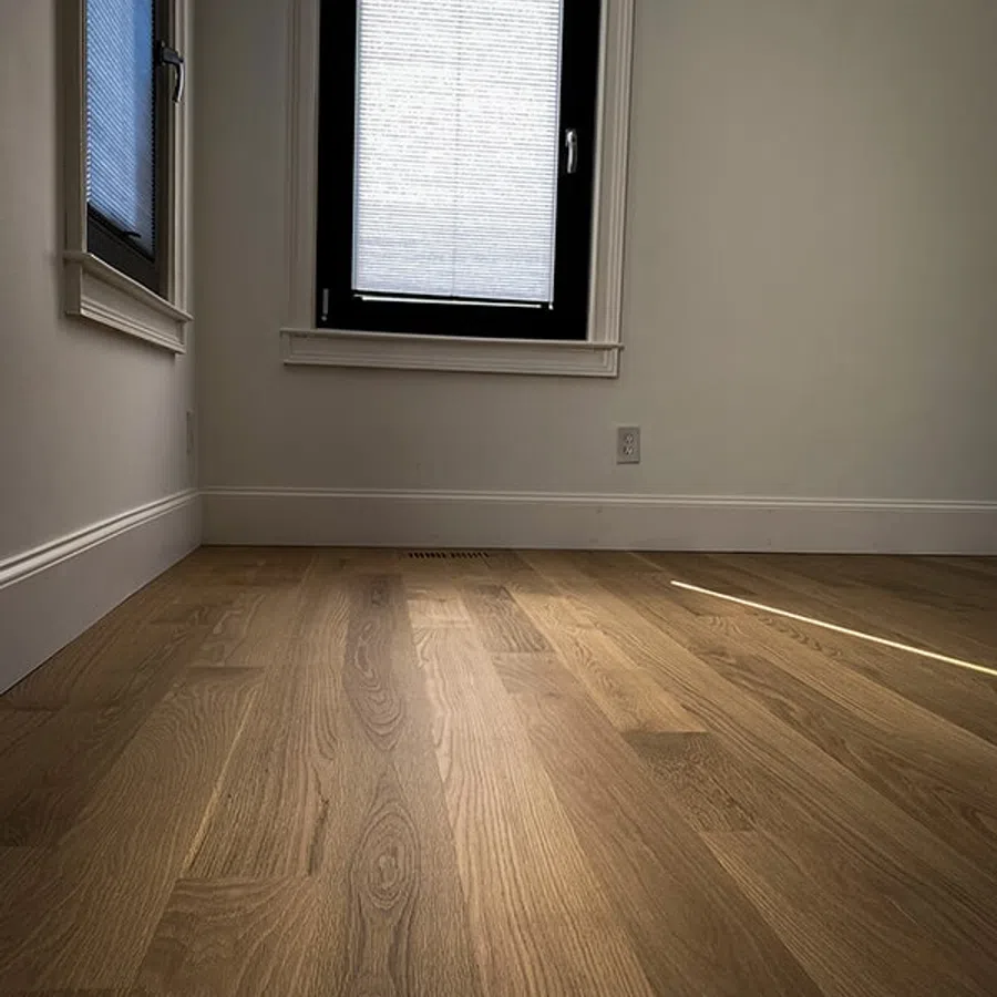 Empty room with wooden flooring, white walls, and two windows with blinds partially closed letting in sunlight.