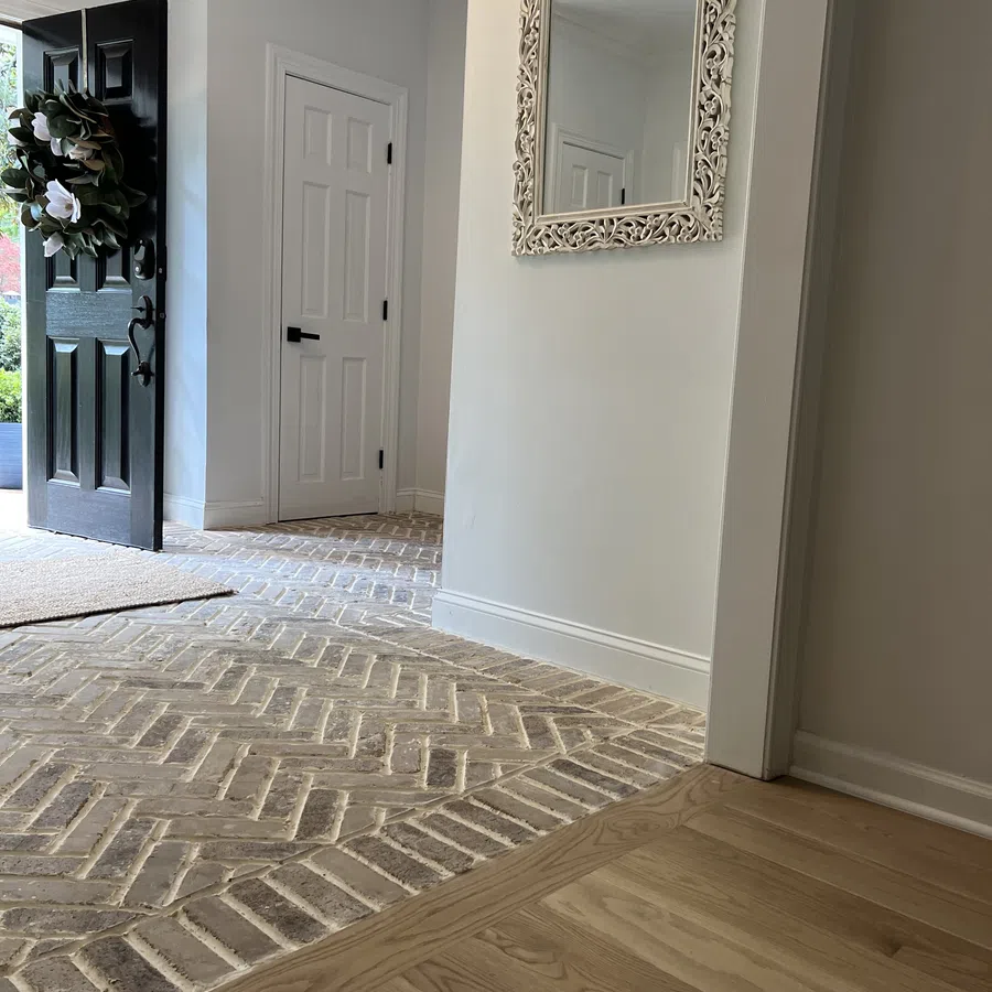 Entryway with open black door, decorative wreath, herringbone brick flooring, white walls, and ornate wall mirror.