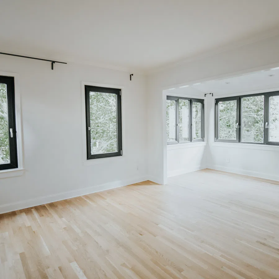 Bright empty room with light hardwood floors and large black-framed windows overlooking green trees.