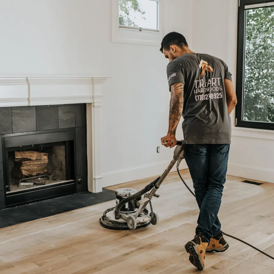 Worker using a floor sanding machine on hardwood floors in a bright room with a fireplace and large window.
