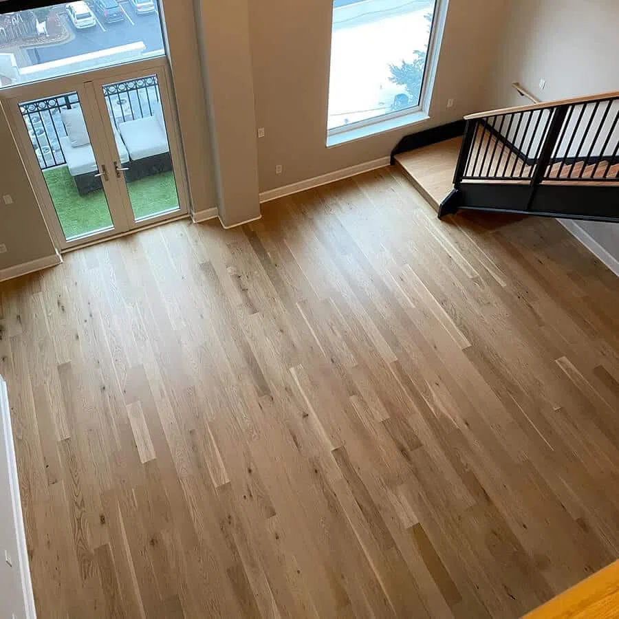 Spacious empty living room with light hardwood floors, large window, glass doors, and metal staircase.