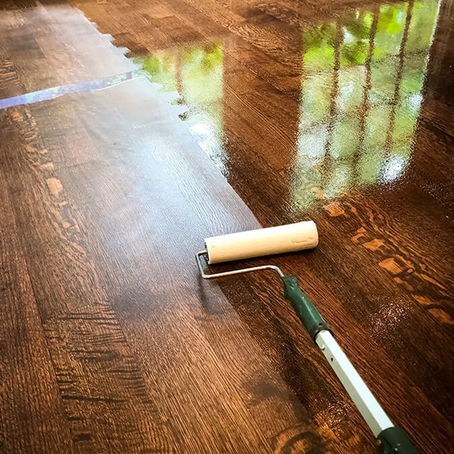 Wood floor being refinished with a paint roller showing wet and dry areas and window reflection.