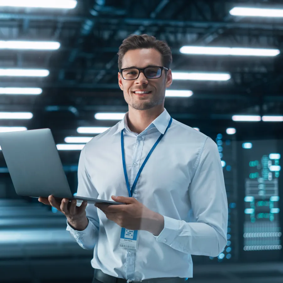 Smiling man in a modern data center holds a laptop and poses confidently for the camera.