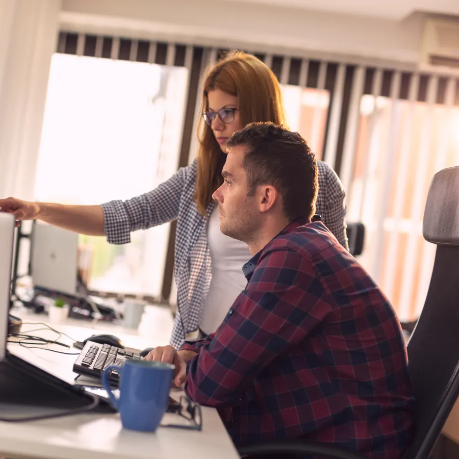 Two colleagues collaborating at a desk with computers in a bright office setting.