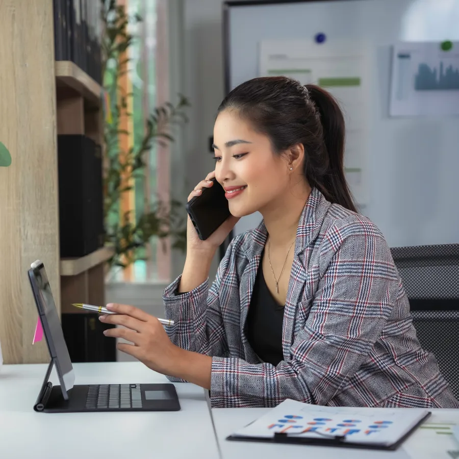 Woman talking on phone while working on tablet in modern office space with plants and bright natural light.