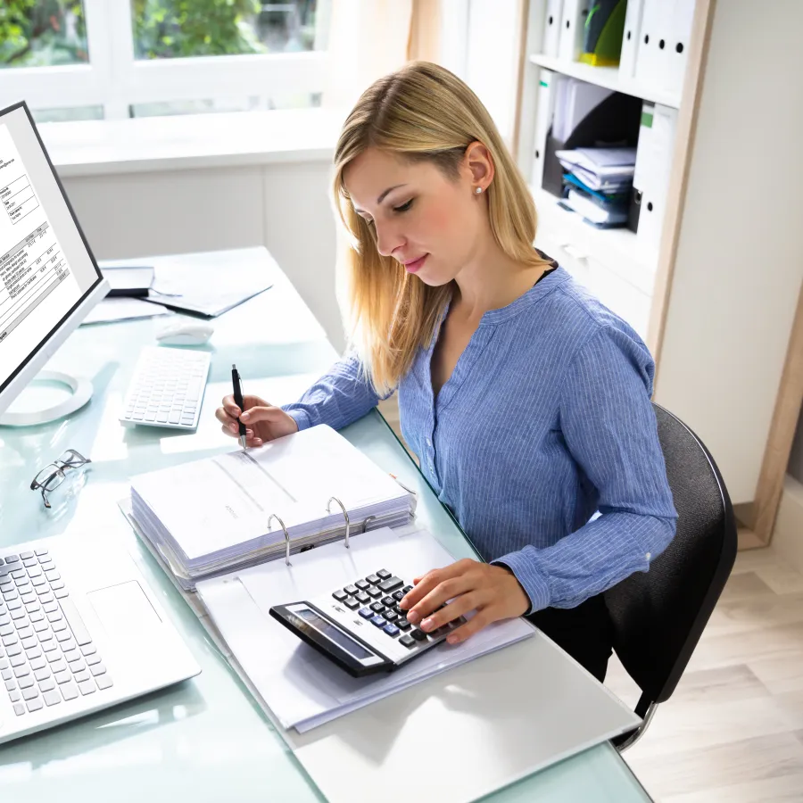 A focused woman works at her desk with a laptop, calculator, and documents in a bright office space.