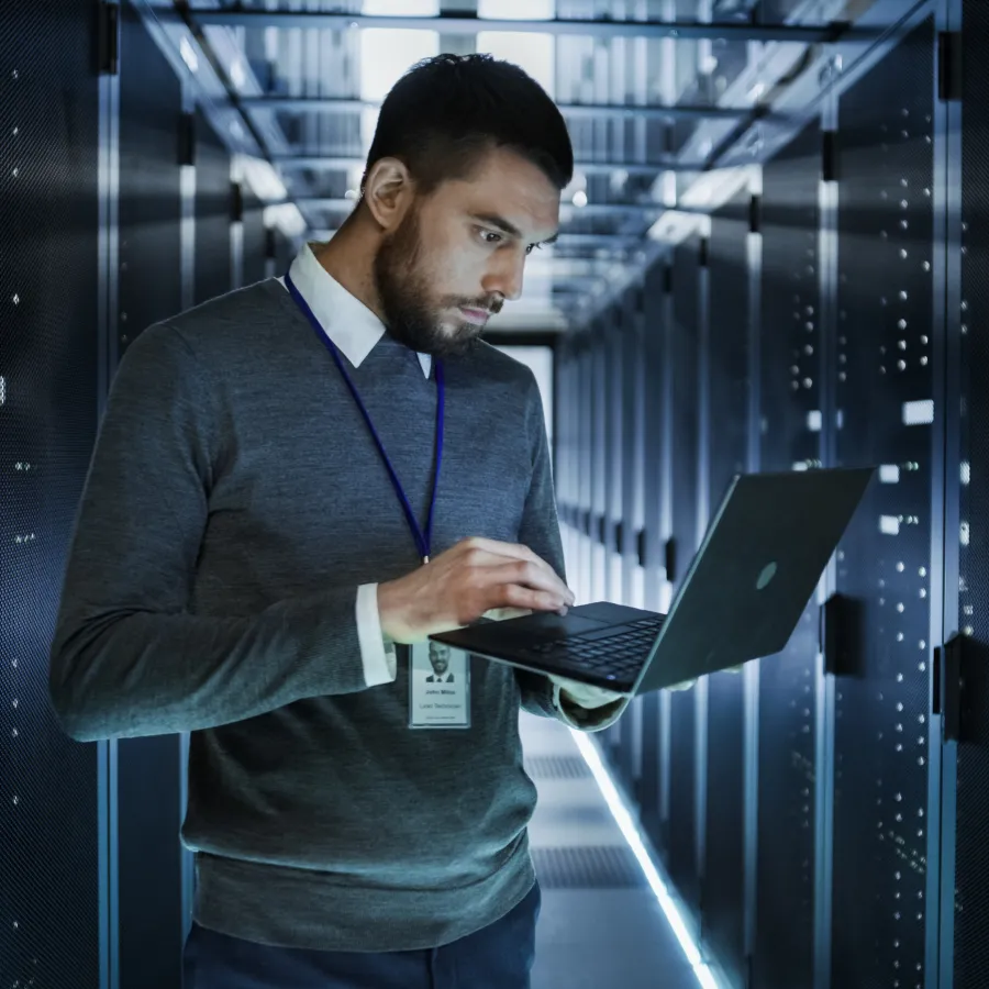 A focused man using a laptop in a data center surrounded by server racks.