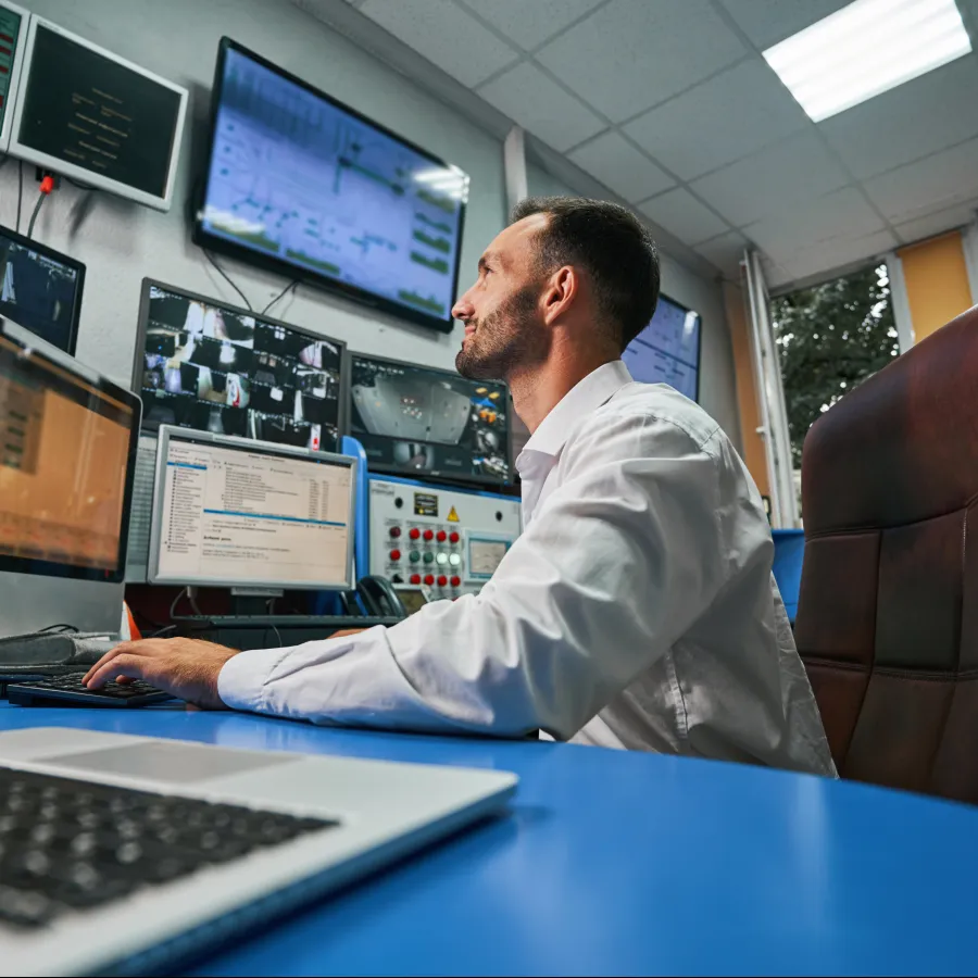 Man working at a computer in a modern control room with multiple screens displaying data.