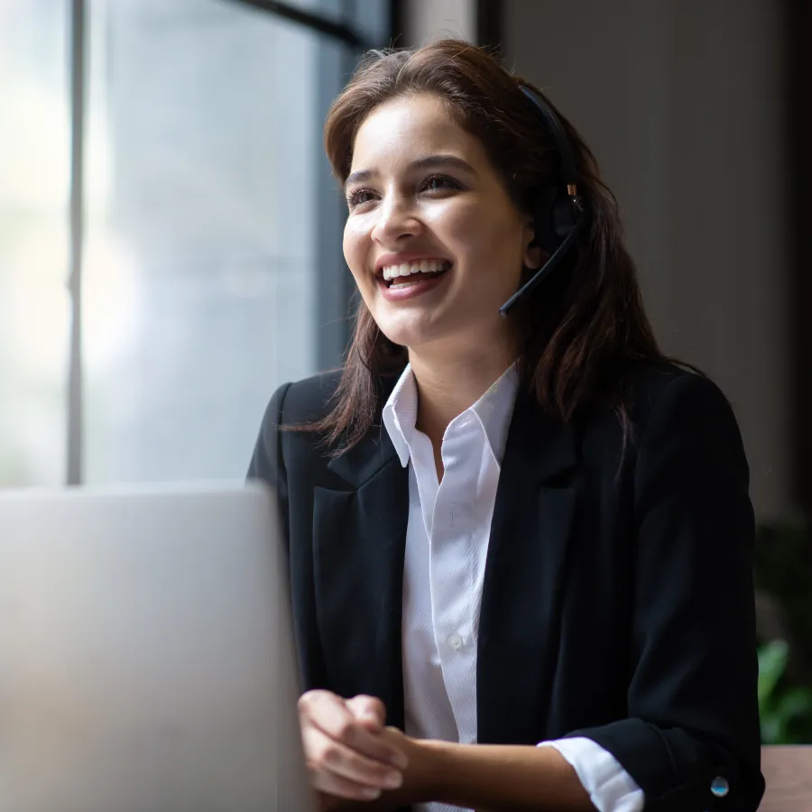 Cheerful woman in a headset working on a laptop in a bright office setting.