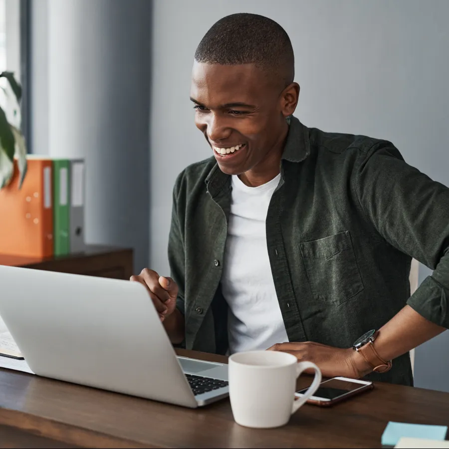 A man smiling while working on a laptop at a desk, with plants and a cup nearby.