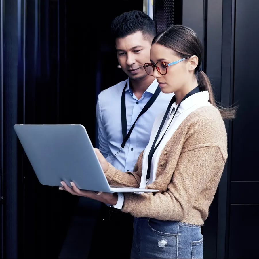 Two IT professionals analyzing data on a laptop in a server room.
