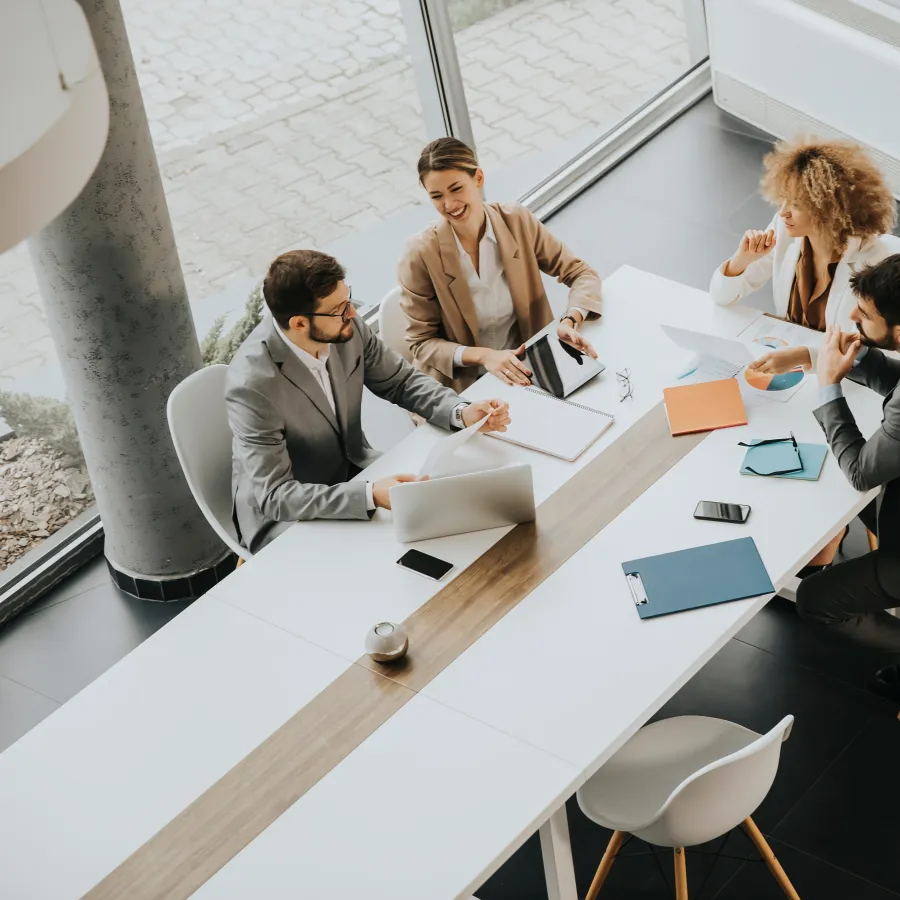 Four professionals in a modern meeting room discussing plans over a table with laptops and documents.
