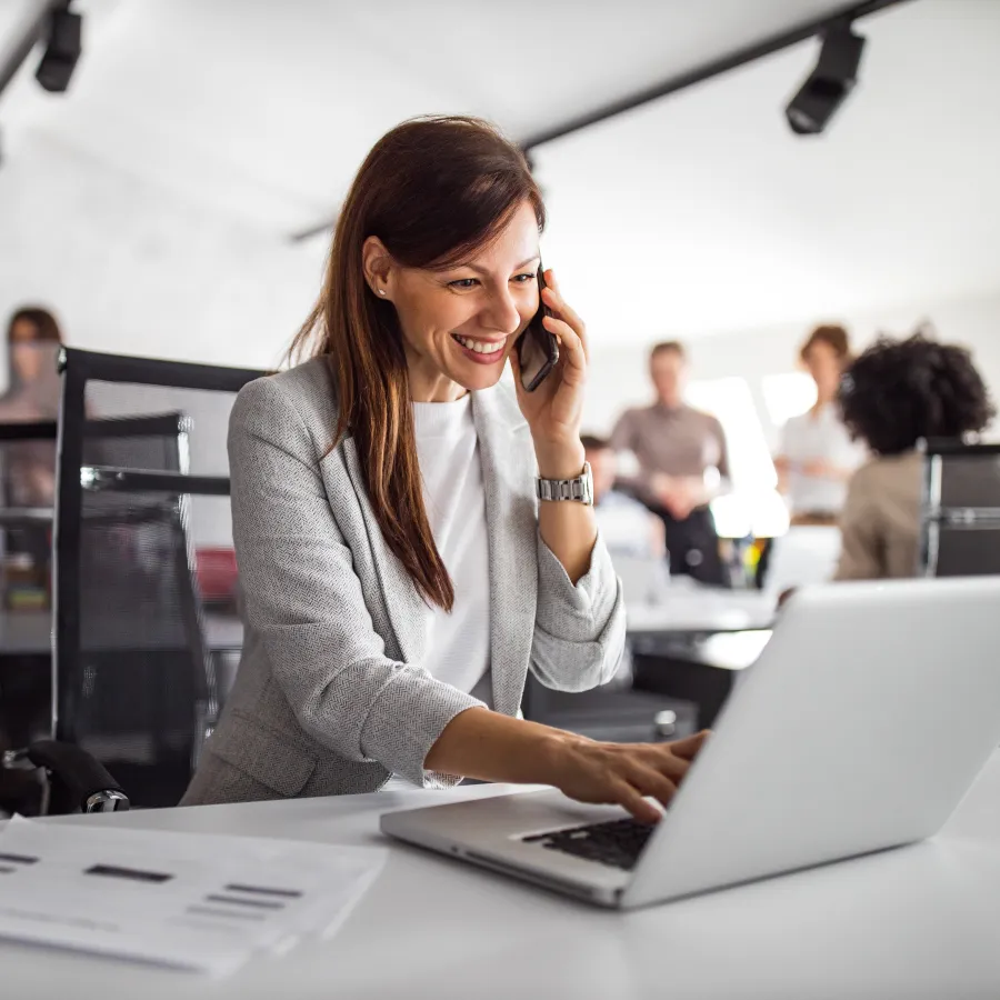 Woman smiling while talking on phone and working on laptop in a modern office setting.