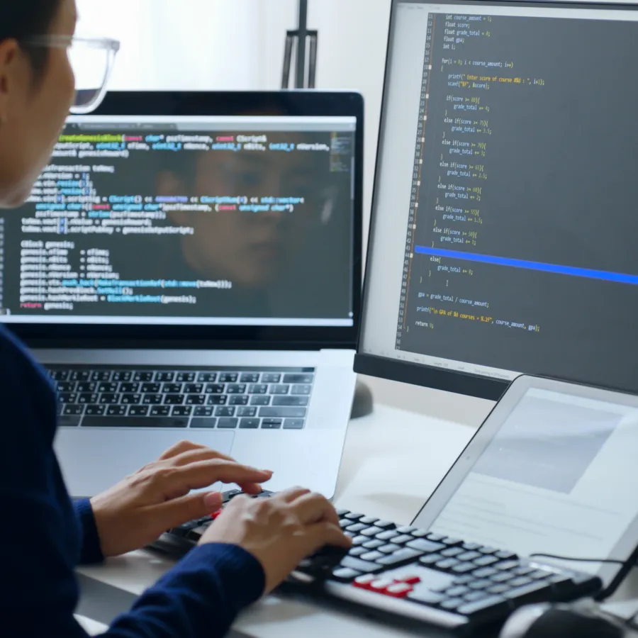 A woman coding on a laptop and monitor with visible lines of code, creating software solutions.