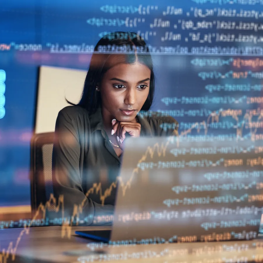 Focused woman analyzing data on a laptop with coding graphics overlay in a modern office.