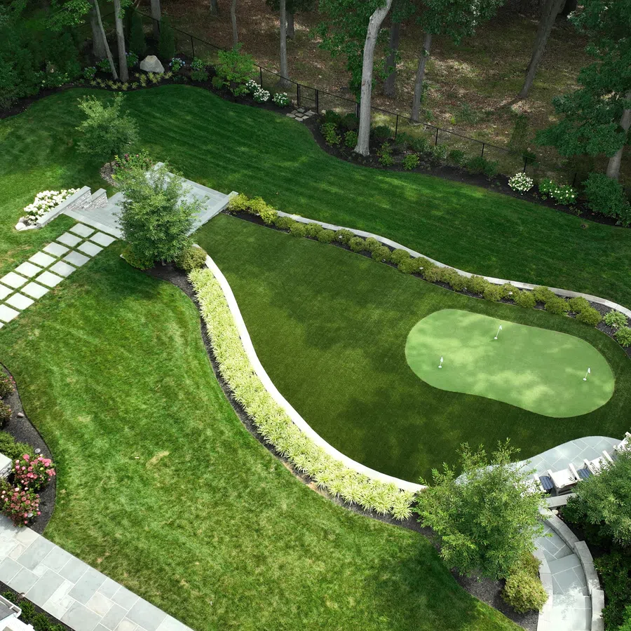 Aerial view of a manicured backyard with a putting green, stone patio, curved pathway, and garden shed.