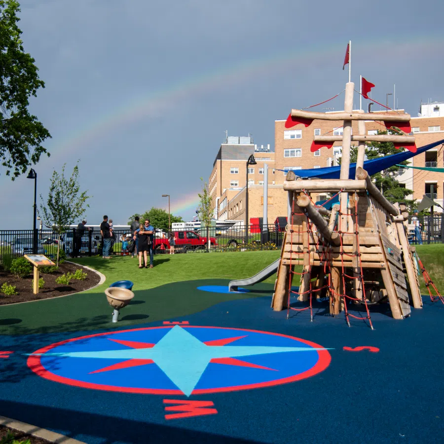 Children's playground with wooden climbing structure and compass design flooring under a faint rainbow in urban park