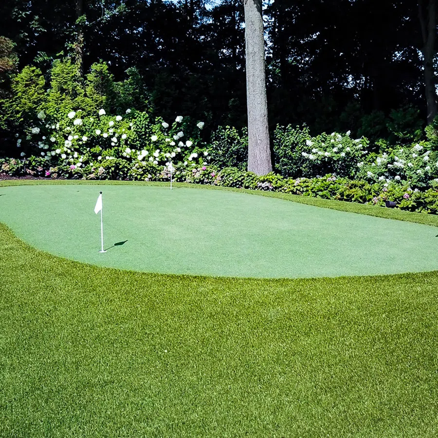 Private backyard golf putting green surrounded by lush grass and flowering plants with trees in background