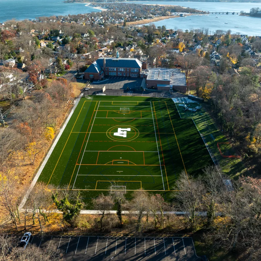 Aerial view of a school with a large sports field, nearby houses, trees, and waterfront in the background.