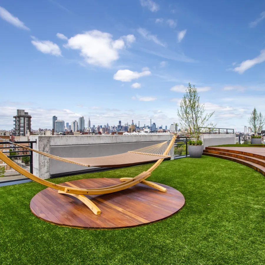 Modern rooftop terrace with green artificial grass, wooden hammock, potted plants, and city skyline under blue sky.