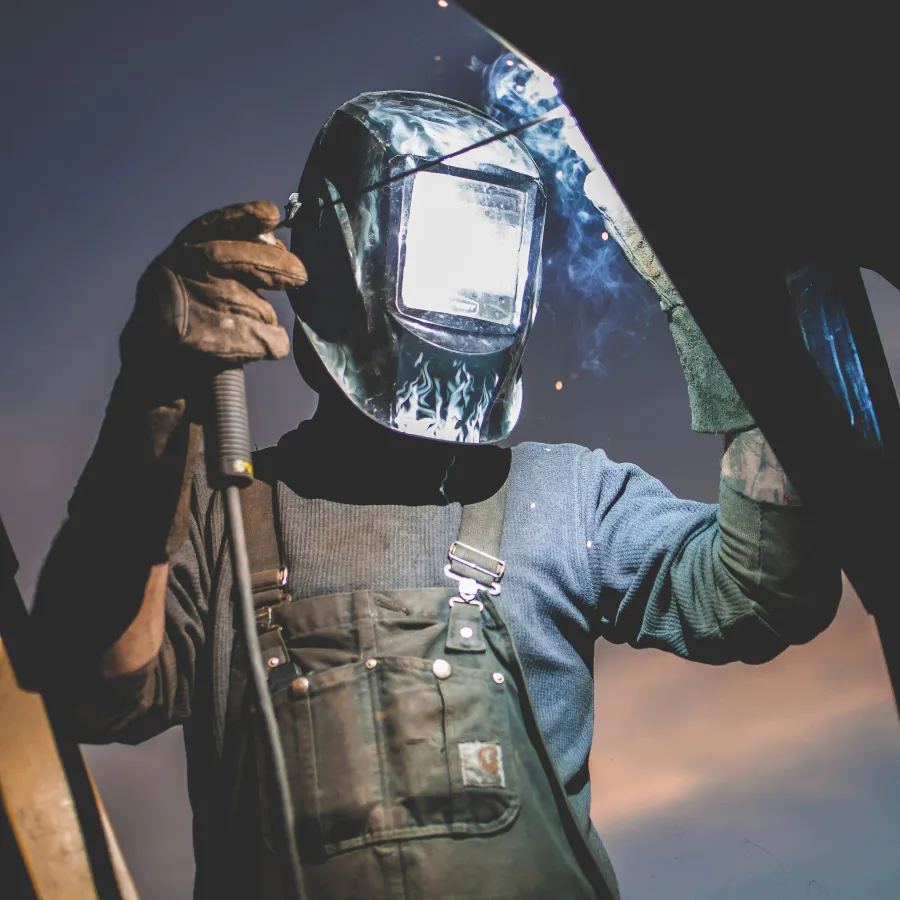 Welder in protective gear welding metal with sparks and smoke against a twilight sky background.