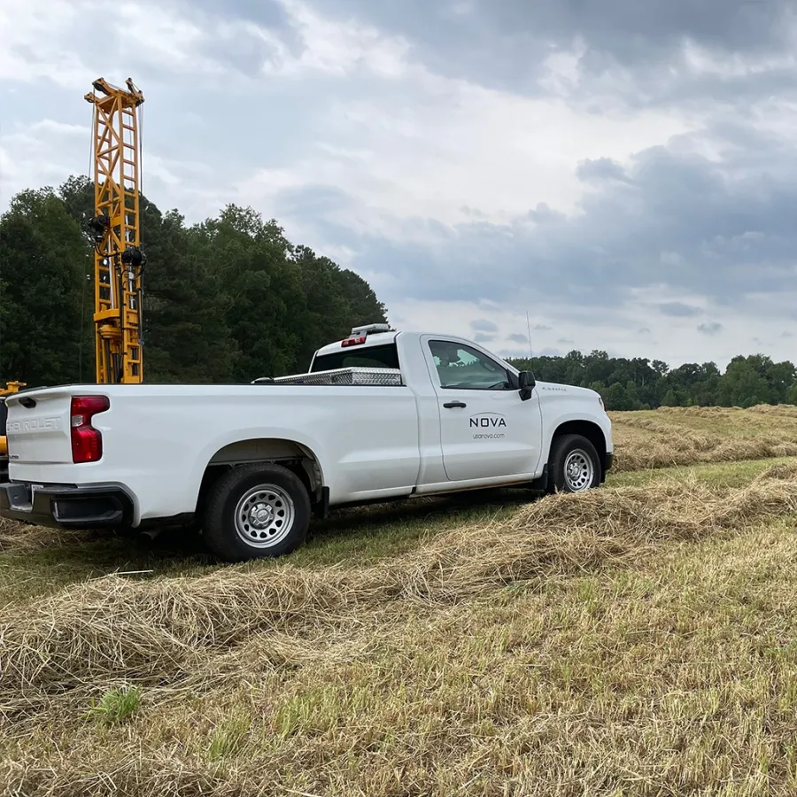 White pickup truck with NOVA logo parked on grassy field near drilling equipment under cloudy sky