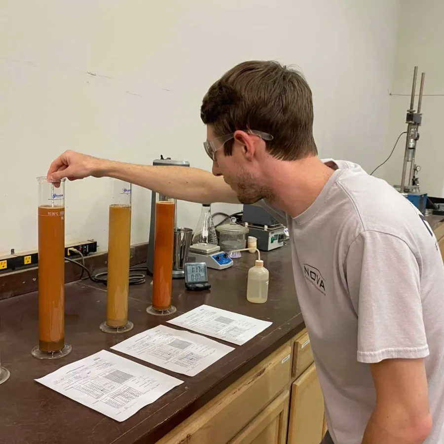 Man wearing safety glasses measuring liquid samples in graduated cylinders on lab counter with documents
