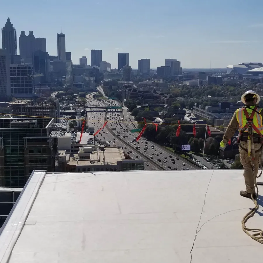 Construction worker with safety harness on rooftop overlooking a busy highway and city skyline under clear sky.