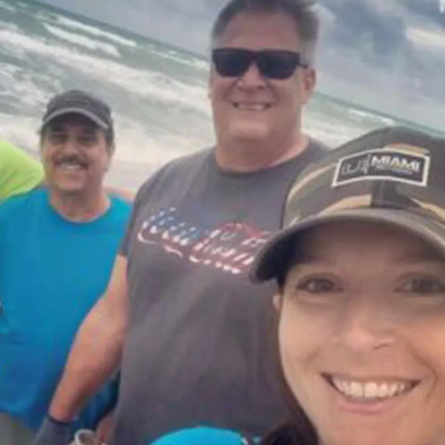 Group of four smiling adults wearing casual clothes and sunglasses at a beach with waves in the background.