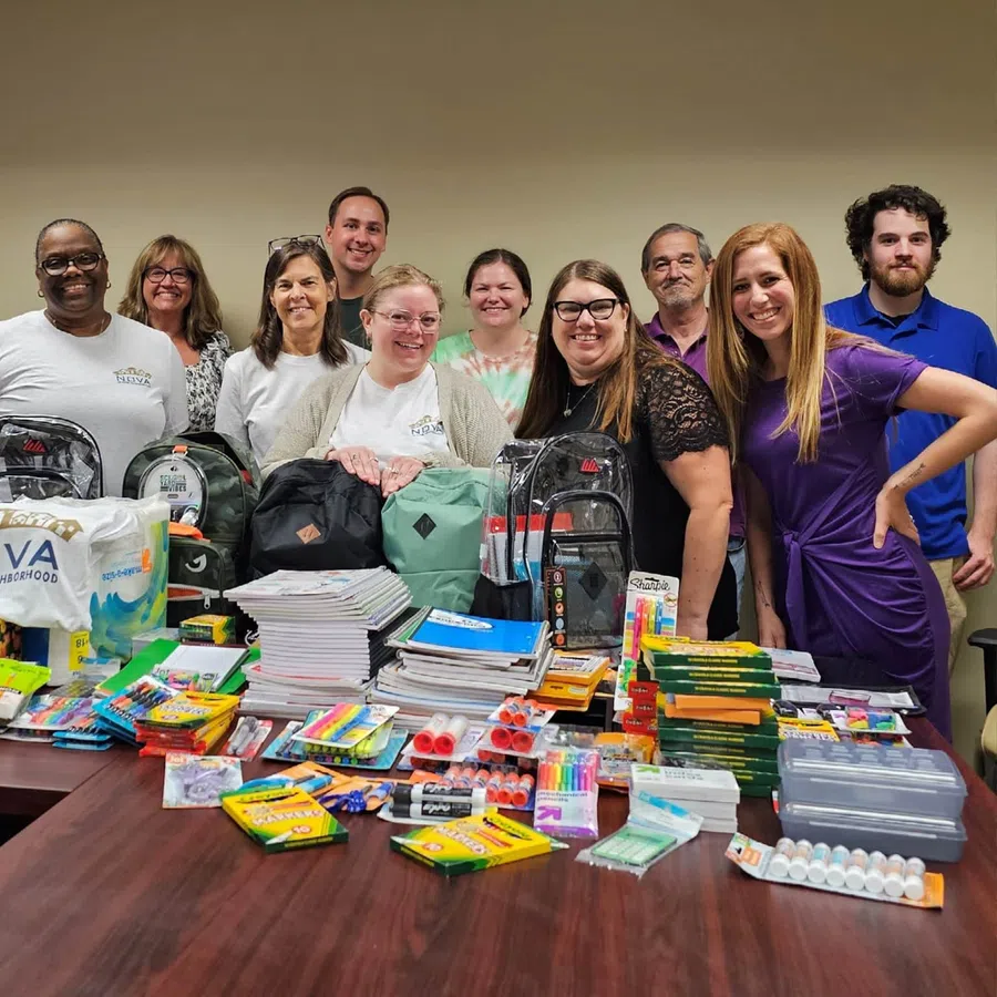 Group of volunteers standing behind a table filled with backpacks and assorted school supplies for donation.