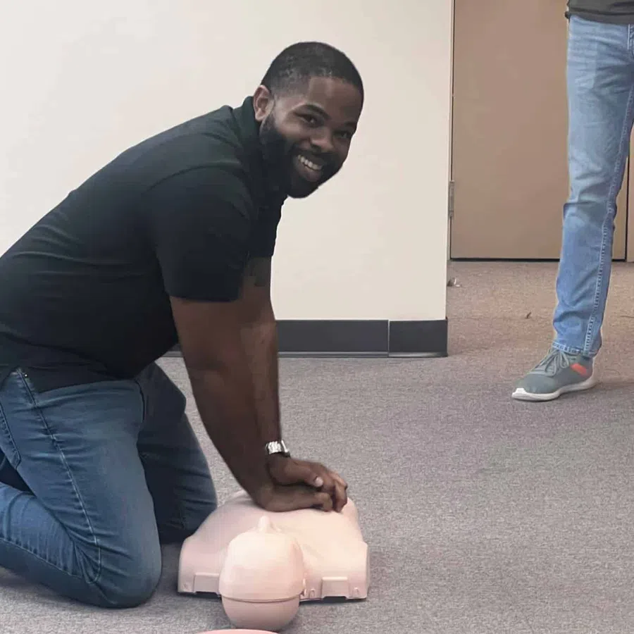 Man kneeling on the floor performing CPR on a training mannequin in an indoor setting.