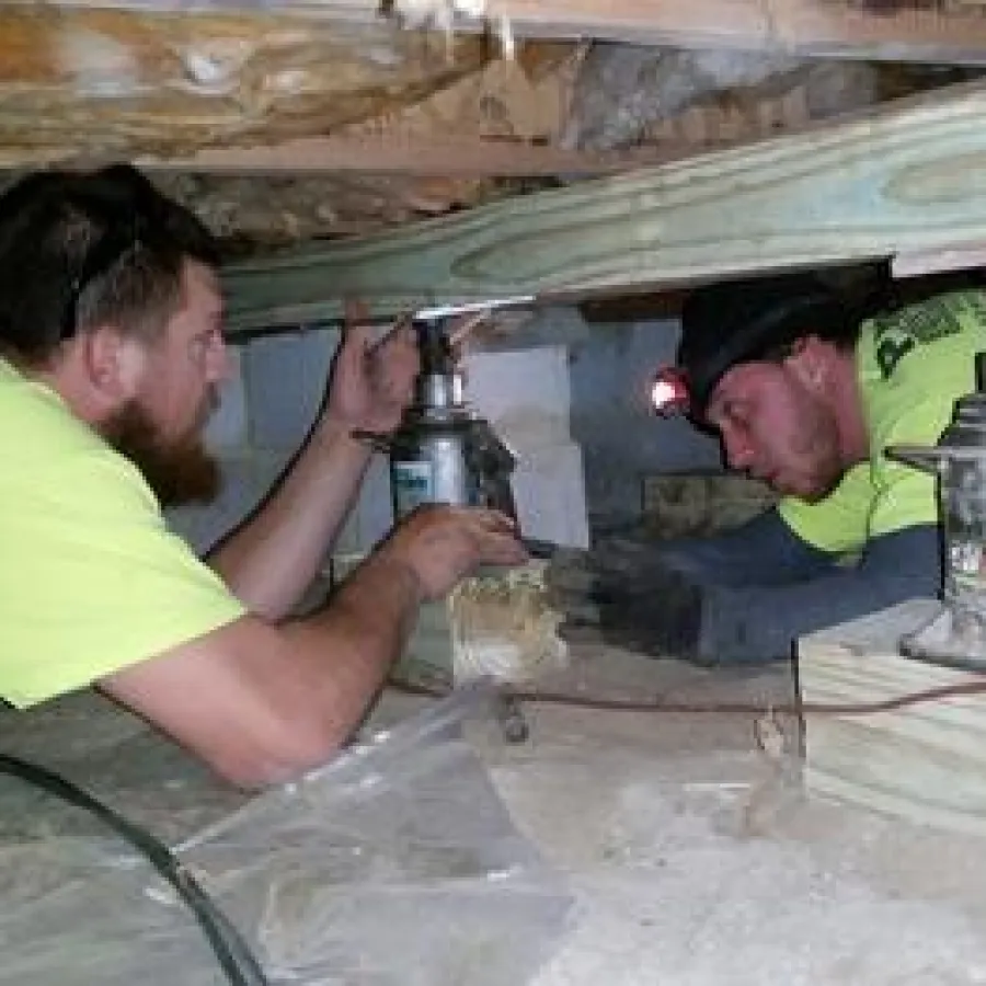 Two workers in yellow shirts inspecting and adjusting foundation jacks under a house crawl space.