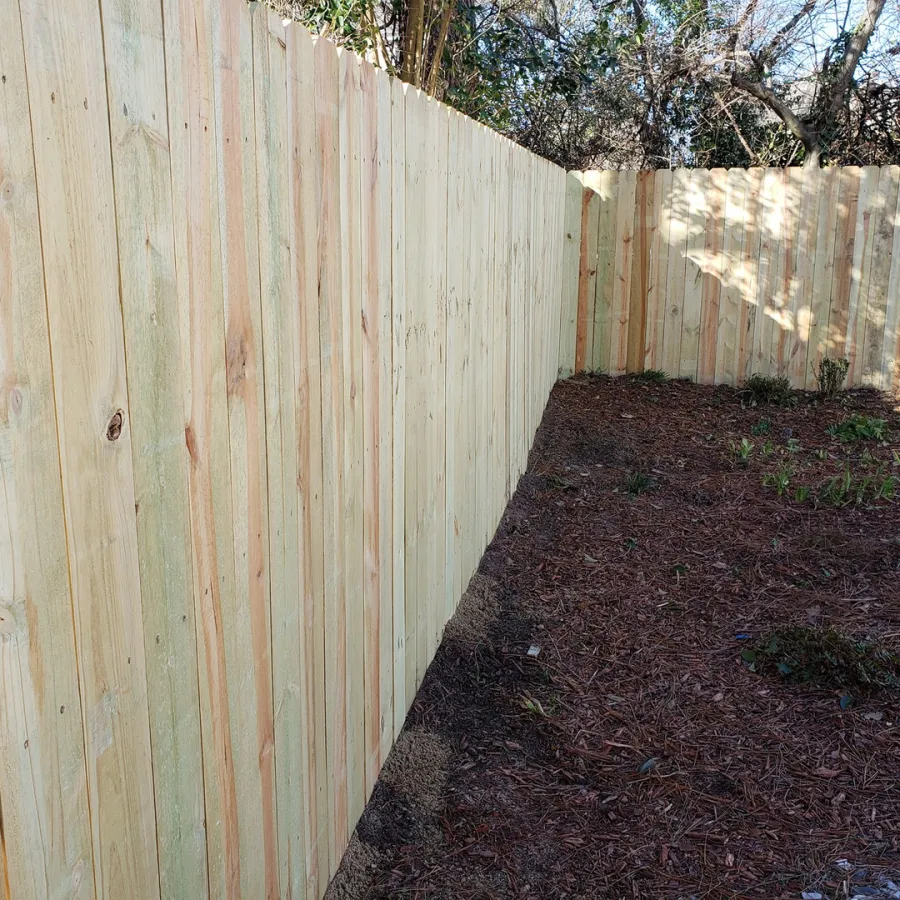 Newly installed wooden fence enclosing a garden area with soil and sparse plants under sunlight.
