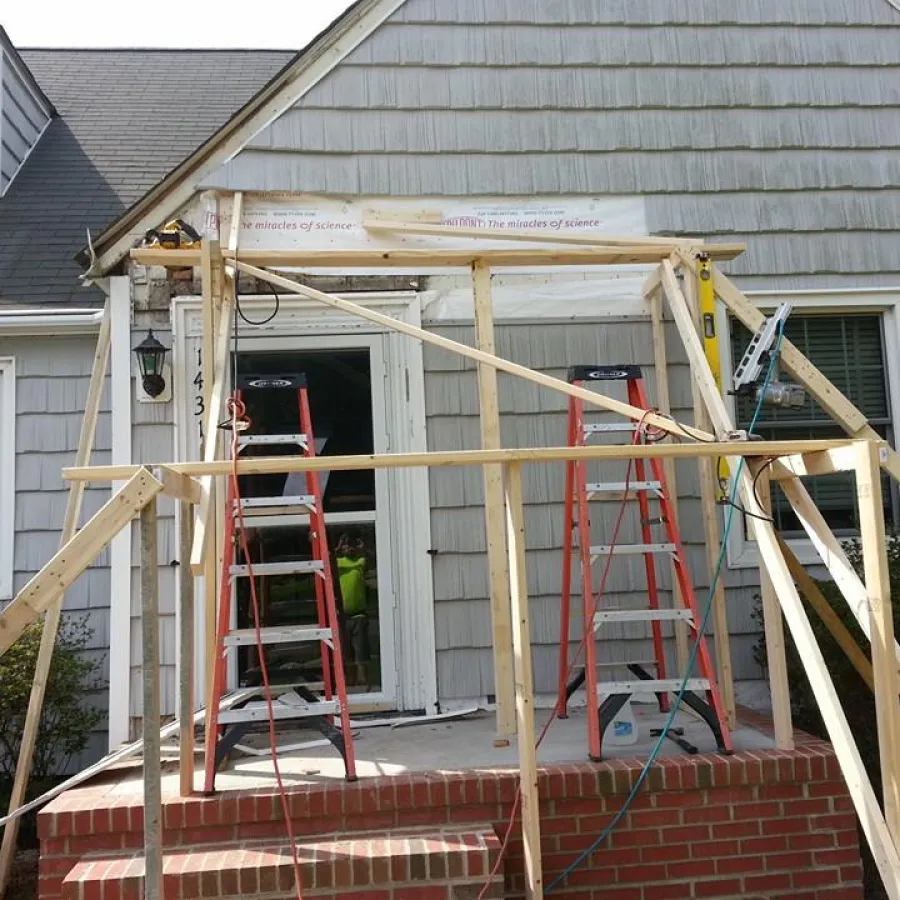 Wooden frame structure being built on a brick porch in front of a gray house with ladders and tools.