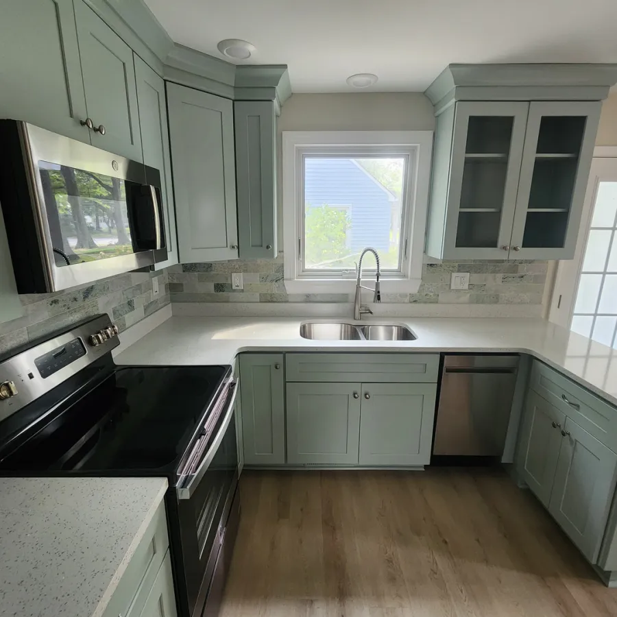 Modern kitchen with light green cabinetry, stainless steel appliances, white countertops, and wood flooring under natural light