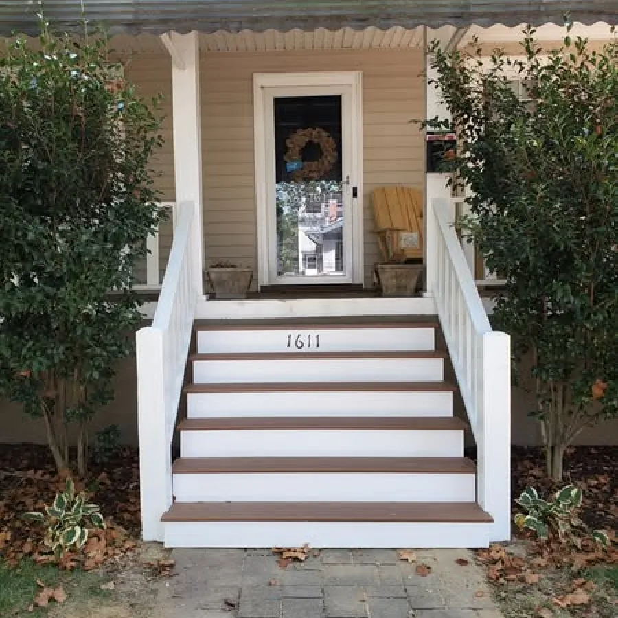 Front porch with white steps, brown treads, white railings, flanked by green shrubs and autumn leaves on ground.