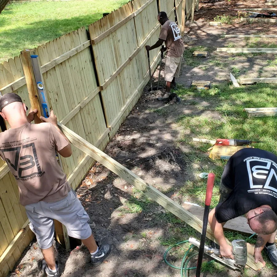 Three workers install a wooden fence using tools and lumber in a sunny yard with grass and trees.