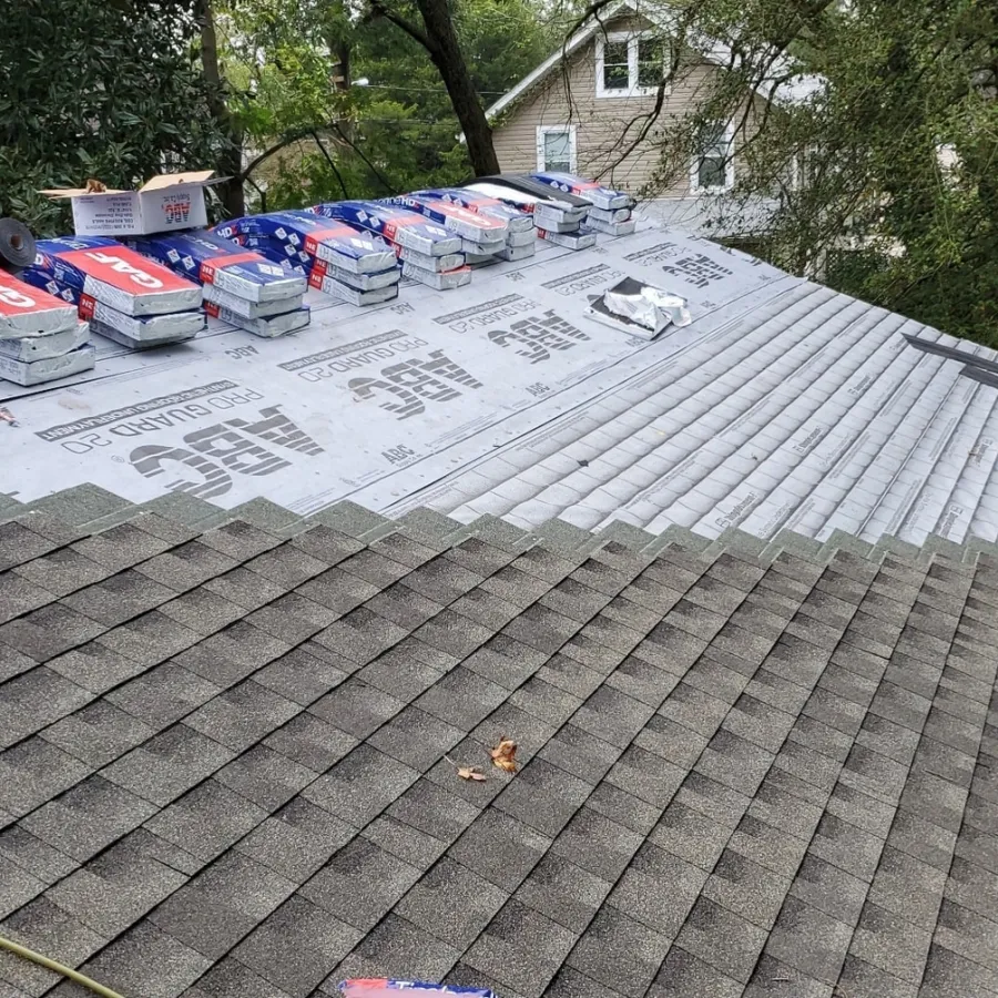 Partially shingled roof with roofing materials and tools ready for installation on a house surrounded by trees.