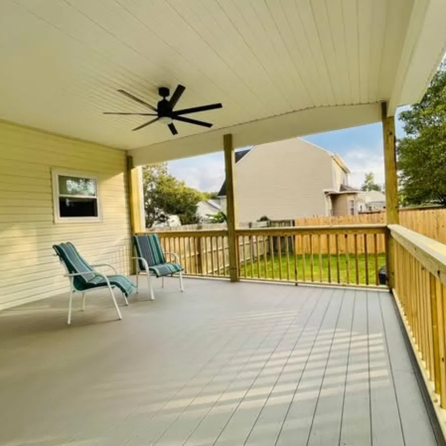 Spacious covered porch with ceiling fan, two green chairs, and wooden railing overlooking backyard.