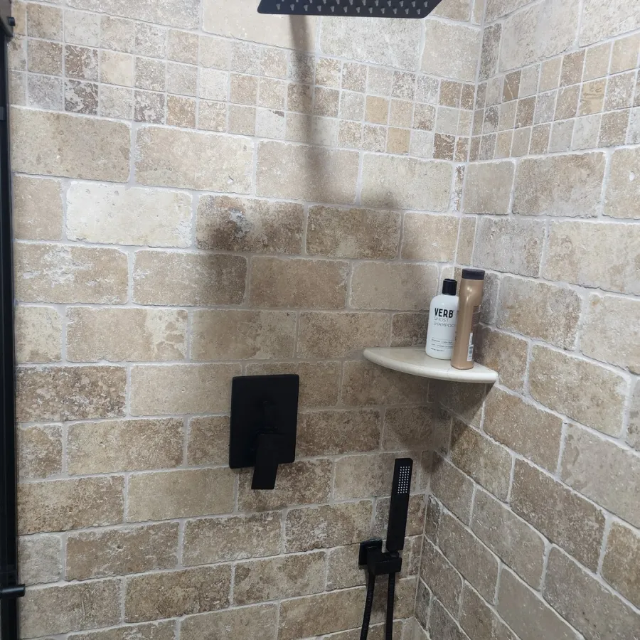 Modern shower with matte black fixtures, beige stone tile walls, and shampoo on a small corner shelf.