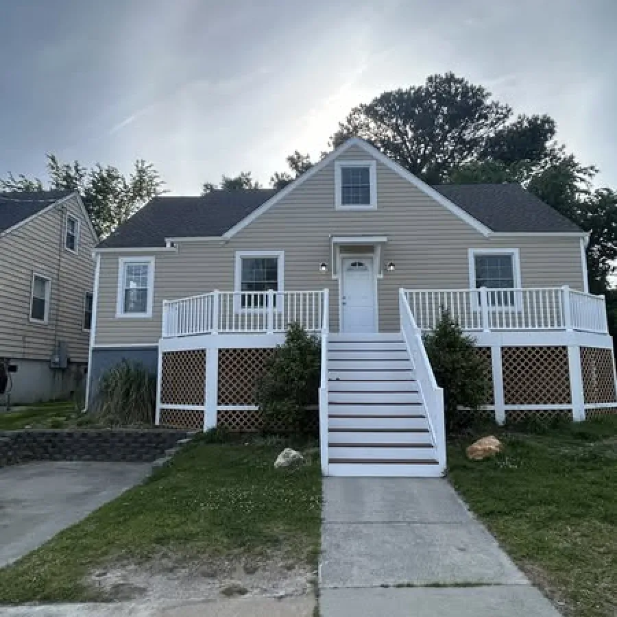 Beige raised house with white railings and stairs surrounded by green lawn under a cloudy sky.