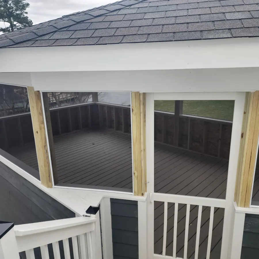 Screened-in porch with white railing and dark wood flooring attached to a house with gray siding and shingled roof.