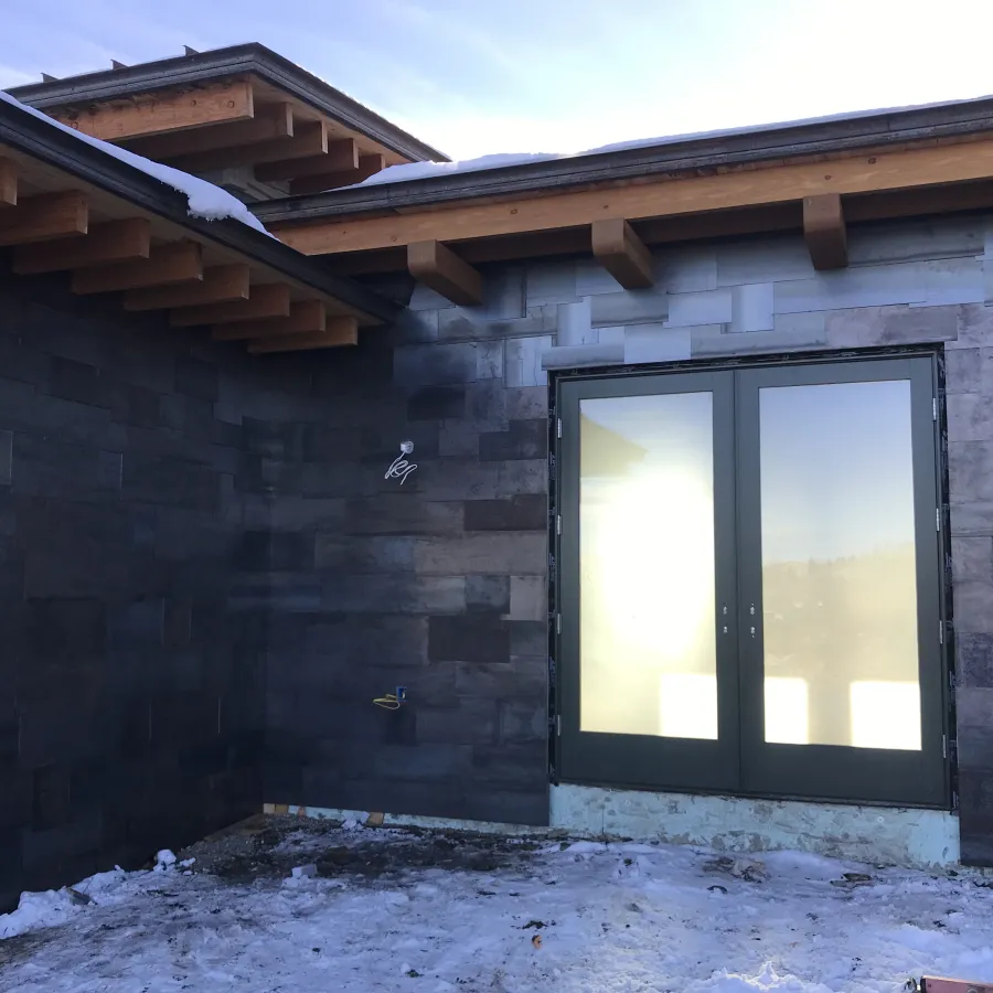 Modern house exterior with dark stone walls, wooden roof beams, snow-covered ground, and frosted glass double doors.